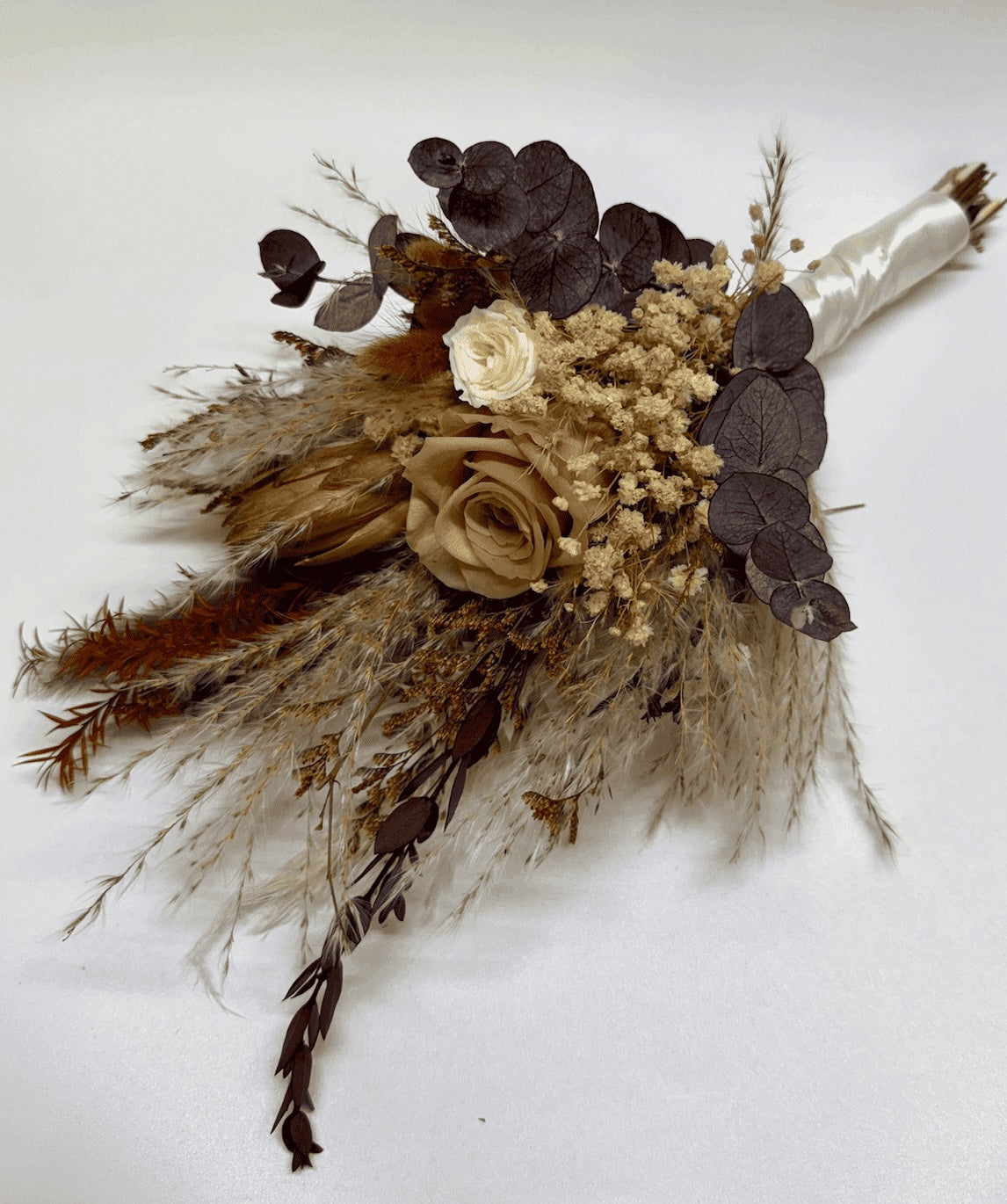 A rustic dried flower bouquet featuring a tan rose, a cream rose, dark burgundy eucalyptus leaves, beige baby's breath, and pampas grass, wrapped in a cream satin ribbon on a white background.