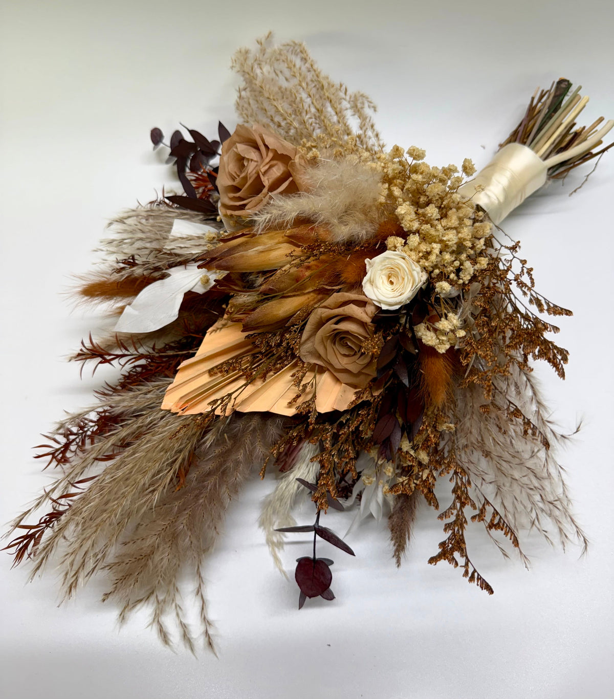 A dried floral bouquet featuring tan and brown paper roses, pampas grass, baby's breath, and a cream ribbon-wrapped handle, photographed on a plain white background.