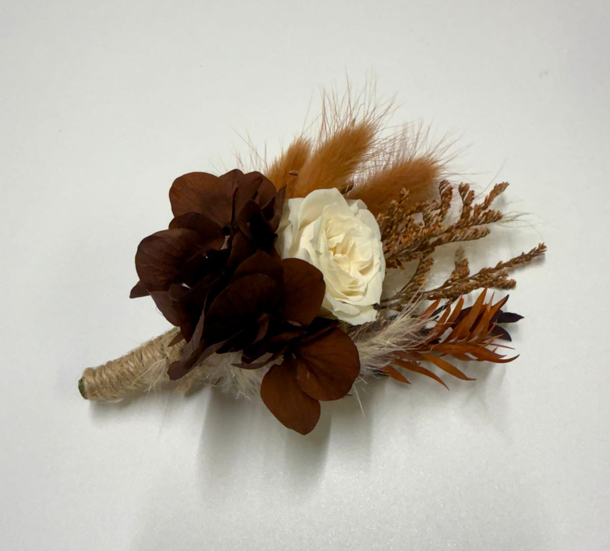 A dried floral boutonniere featuring dark brown hydrangea-like blooms, a cream-colored rose, orange-brown feathers, and natural twine wrapping on a white background.