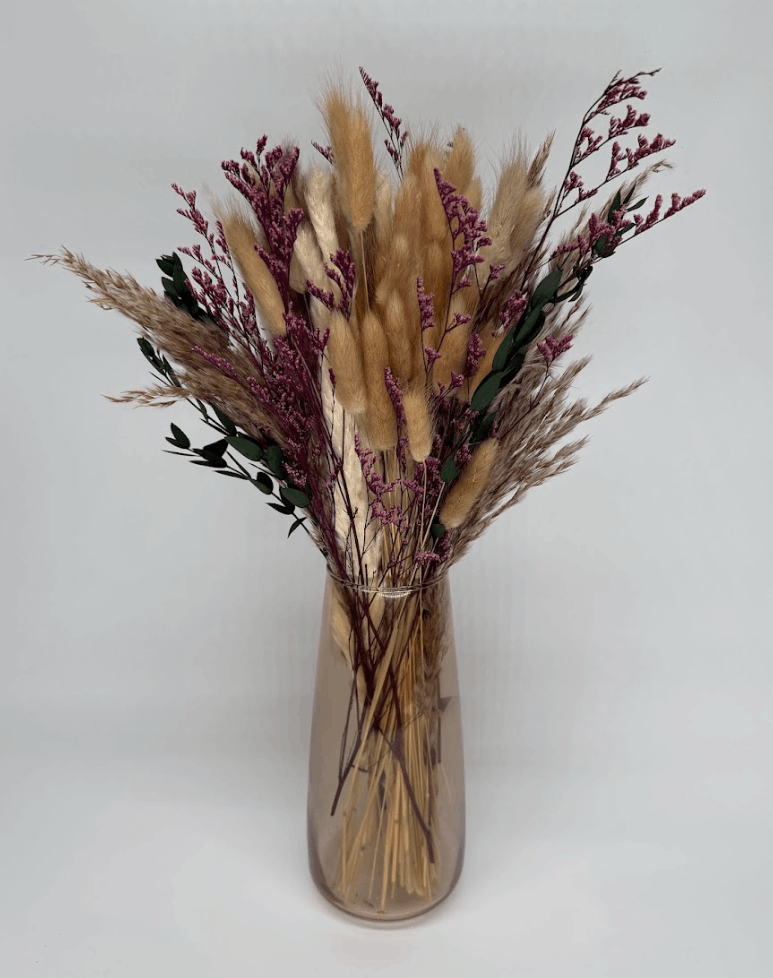 Boho dried flower arrangement with pampas grass, pink stems, and eucalyptus in a clear glass vase on a white background