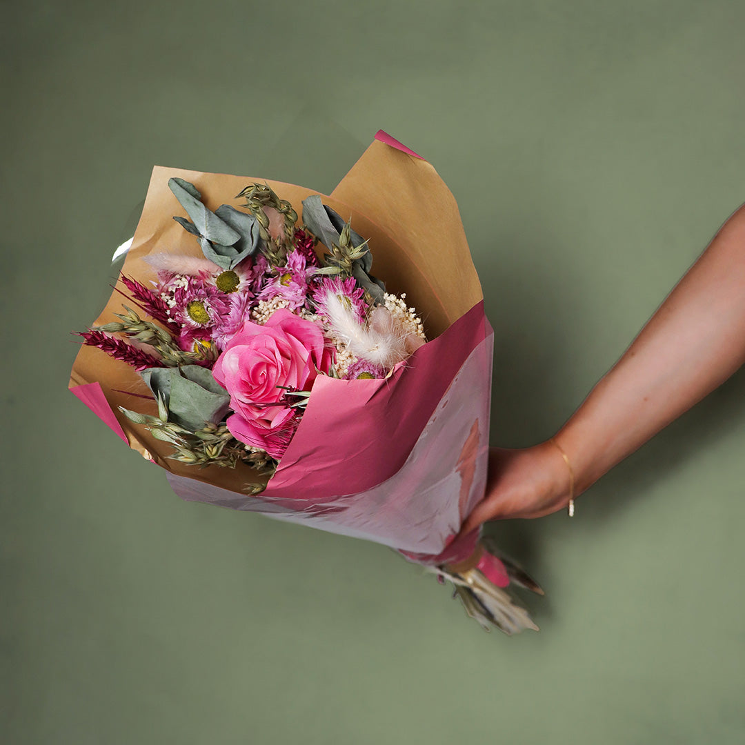 Bouquet of flowers wrapped in brown paper with pink tape held by a person against a green background