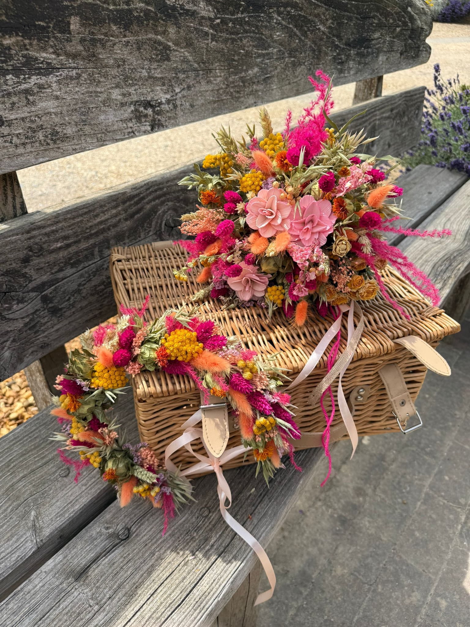 A rustic wicker picnic basket with two floral bouquets in pink, orange, and yellow, tied with pale pink ribbons, resting on a weathered wooden bench.