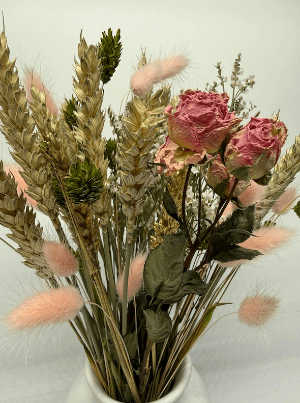 Dried flower arrangement with pink roses, wheat grasses, and bunny tails in a white vase on a light background