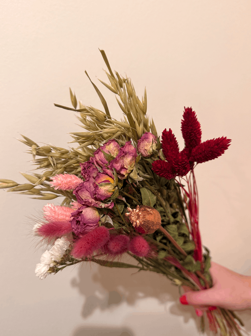 Bouquet of dried flowers with pink, red, and green hues held against a plain background.