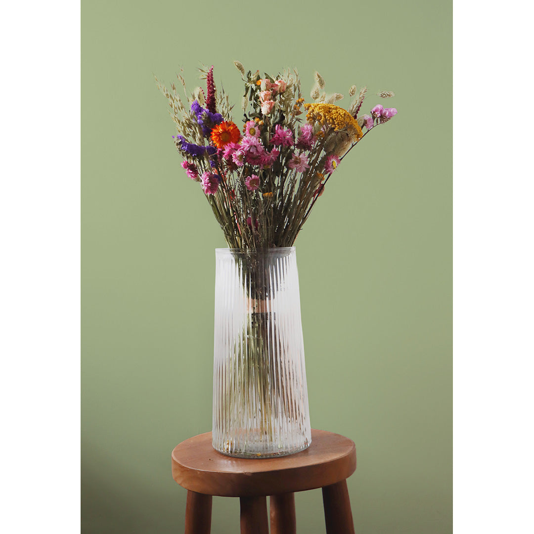 Clear ribbed vase with dried flowers on a wooden stool against a green background