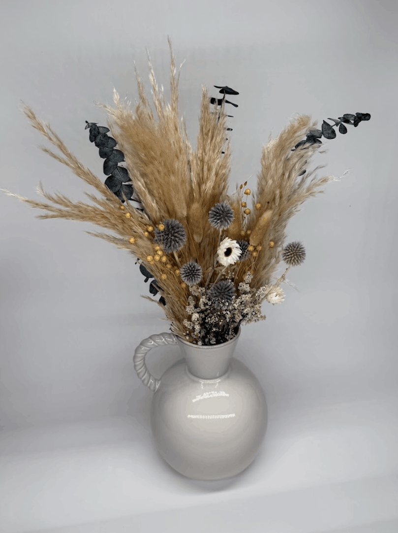 Dried flower bouquet in silver grey and dusky blue tones with pampas grass in a white ceramic vase on a dark background