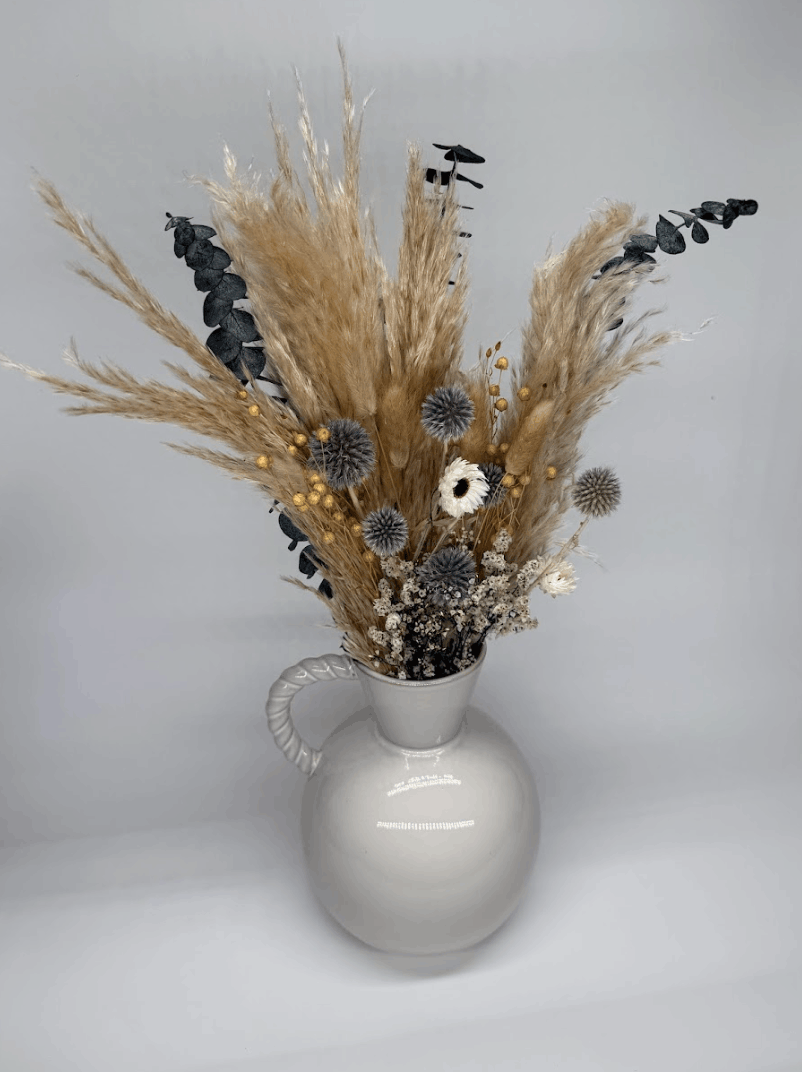 Front view of silver grey dried flower bouquet with pampas grass, blue globe thistles, and eucalyptus in a white ceramic vase on a dark background