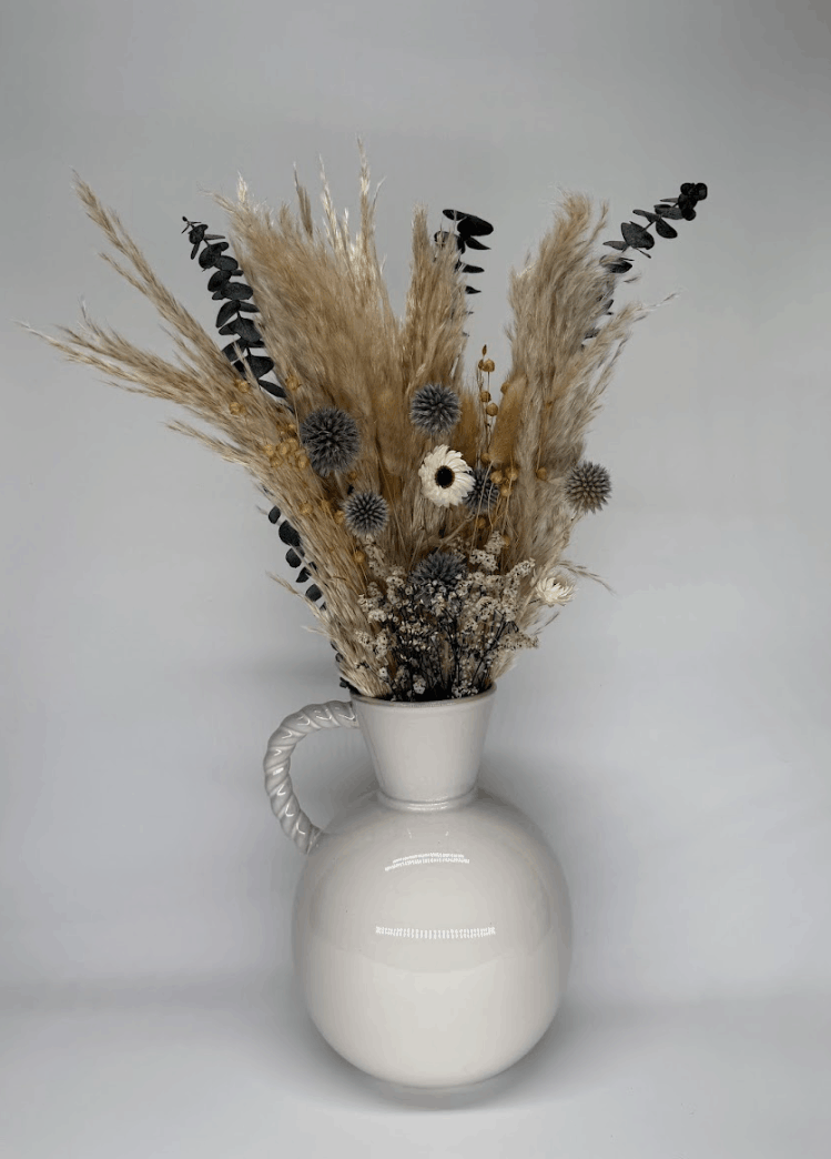 Side angle of silver grey dried flower bouquet with blue globe thistles, pampas grass, and white dried daisy in a white ceramic vase