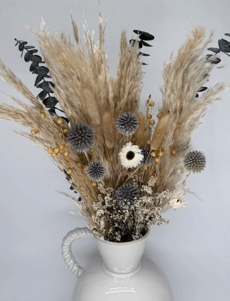 Silver grey and dusky blue dried flower bouquet with pampas grass, globe thistles, and eucalyptus in a white ceramic vase on a light background