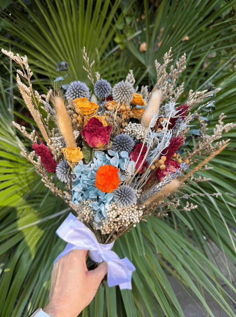 A hand holding a dried flower bouquet with orange, red, blue, and beige botanicals tied with a light purple ribbon, set against a background of green palm leaves.