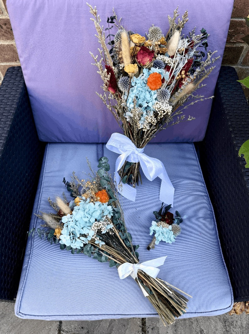 A dried flower bouquet and matching boutonniere set displayed on a lavender cushioned chair outdoors. The floral arrangement features light blue hydrangeas, orange poppy-like blooms, eucalyptus, and pampas grass tied with white ribbons.
