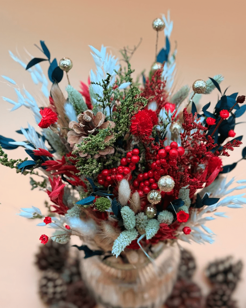 Decorative floral arrangement with red flowers, greenery, and pinecones on a beige background