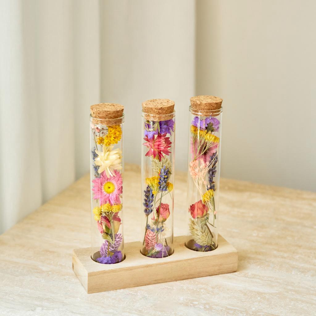 Three glass tubes with cork lids containing dried wildflowers on a wooden stand.