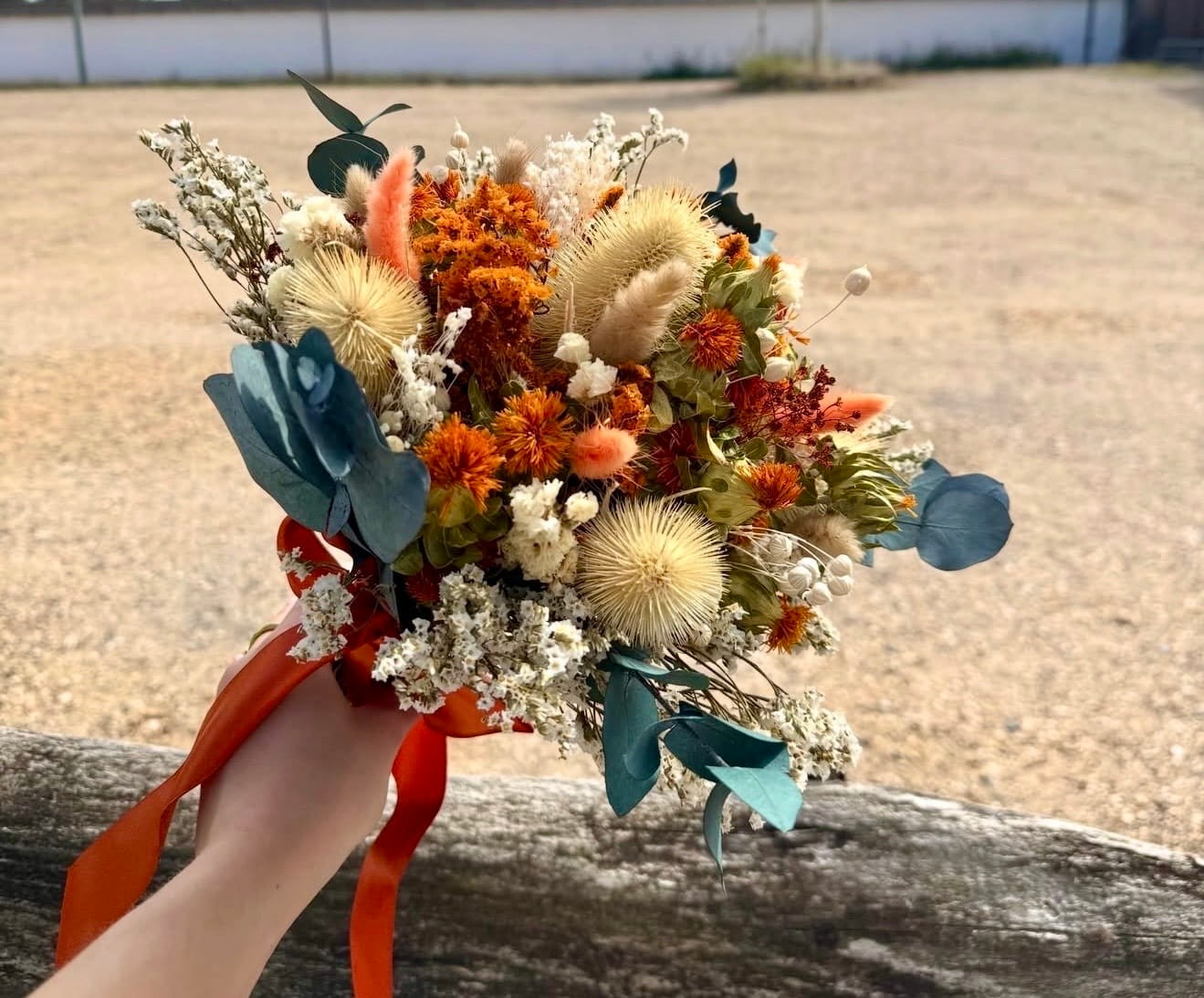 A hand holding a dried flower bouquet with orange, beige, white, and teal botanical elements tied with an orange ribbon, set against a blurred outdoor sandy background.