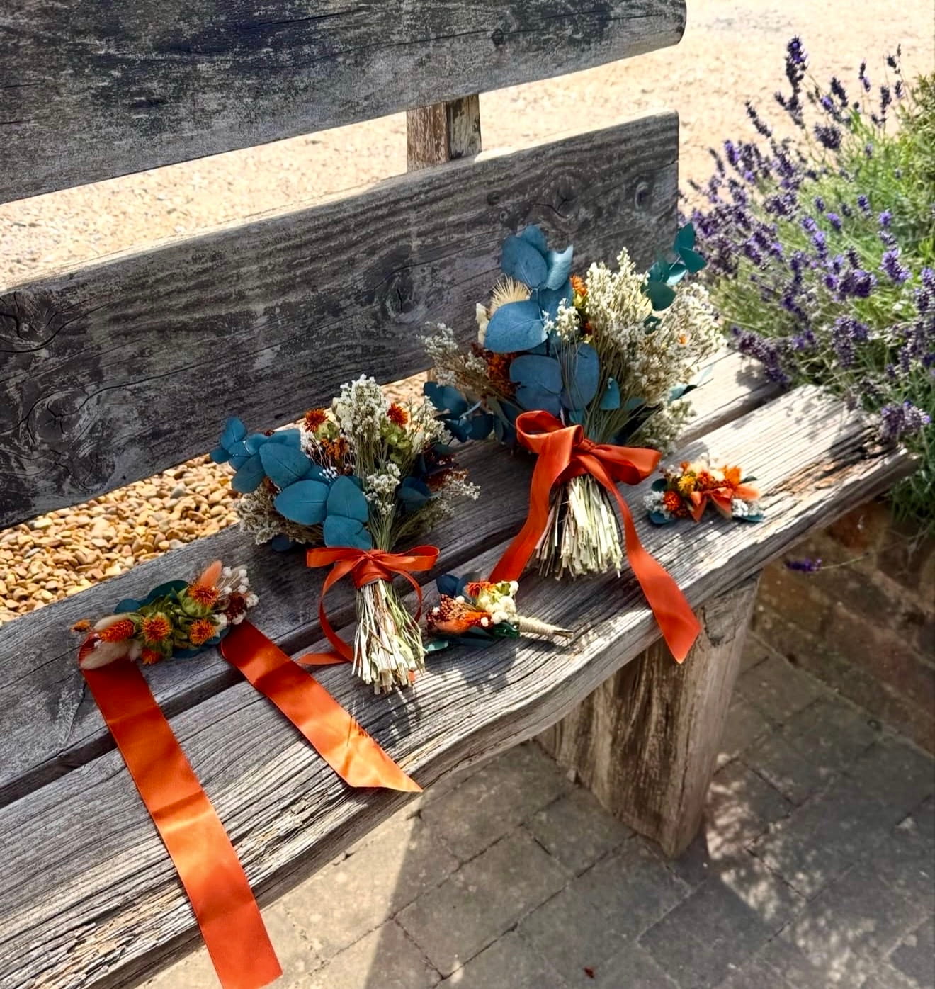 Three small dried floral wristlet bouquets with blue eucalyptus, white baby's breath, and bright orange ribbons are arranged on a rustic wooden bench outdoors next to lavender plants.