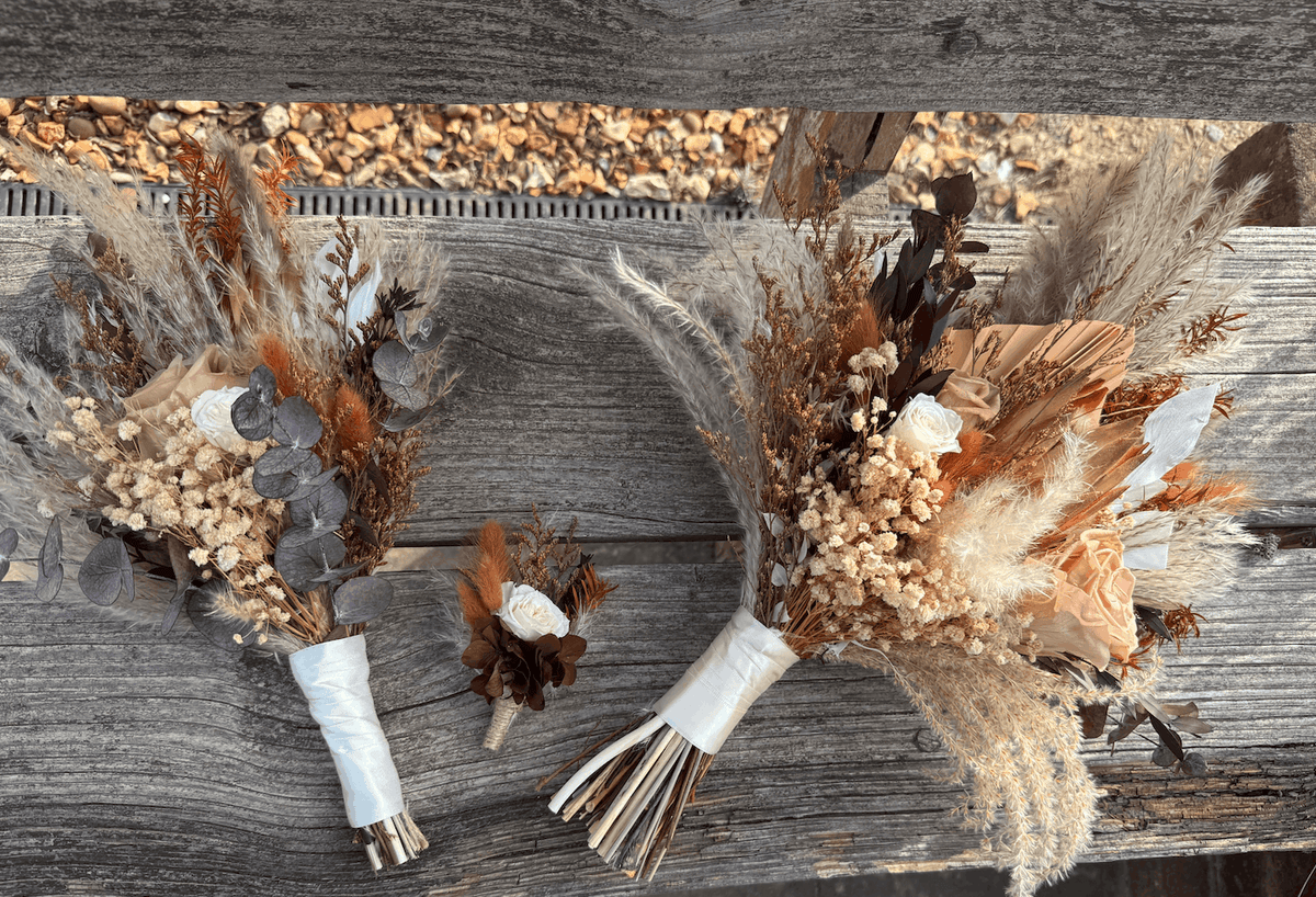 Three dried floral arrangements and a matching boutonniere laid on weathered wooden boards outdoors. The bouquets include beige pampas grass, brown and tan botanicals, muted orange dried flowers, and gray-green eucalyptus leaves, with white ribbon wraps at the stems.