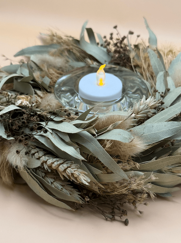Decorative wreath with a small candle inside, surrounded by greenery and feathers on a beige background.