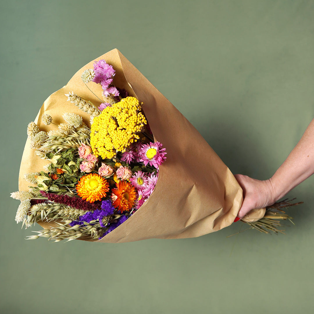 Bouquet of colorful flowers wrapped in brown paper held by a hand against a green background