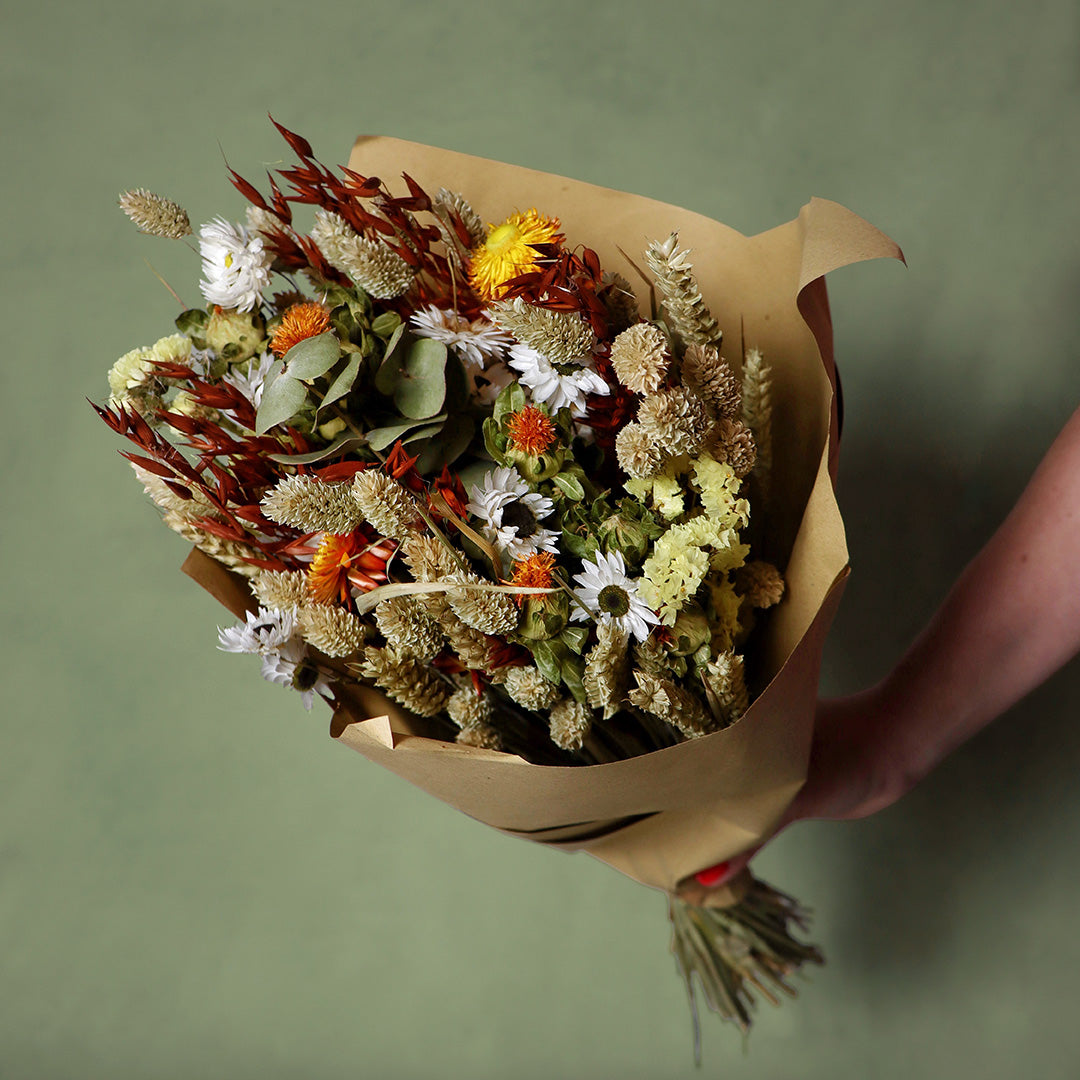 Bouquet of dried flowers wrapped in brown paper held by a hand on a green background