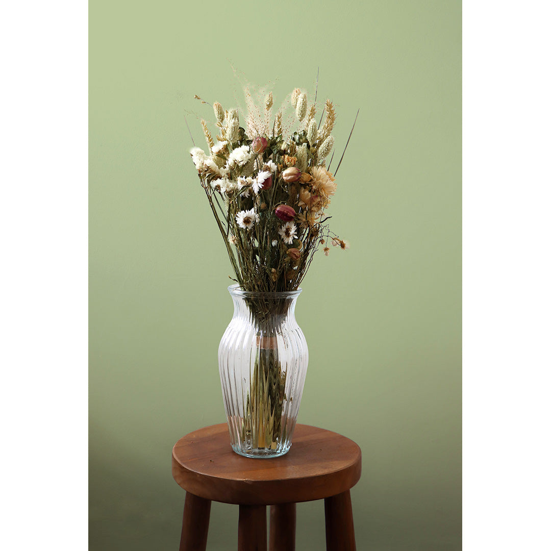 Clear glass vase with dried flowers on a wooden stool against a green wall