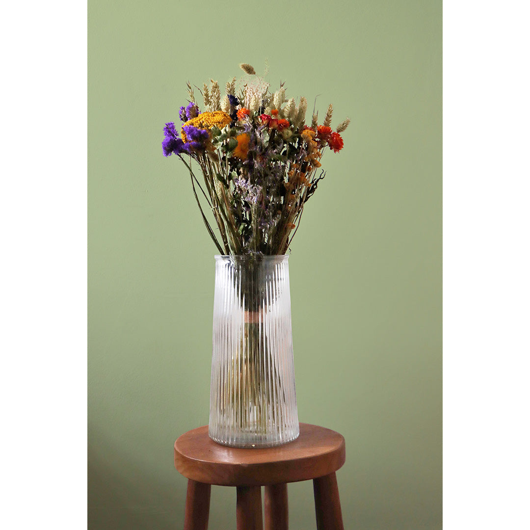 Clear glass vase with dried flowers on a wooden stool against a green wall