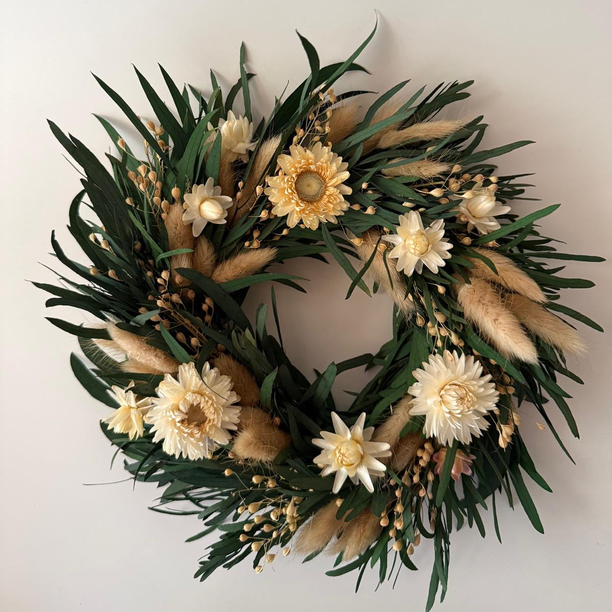 A circular dried flower wreath featuring green foliage, cream and pale yellow flowers, and tan pampas grass, displayed against a plain off-white wall.