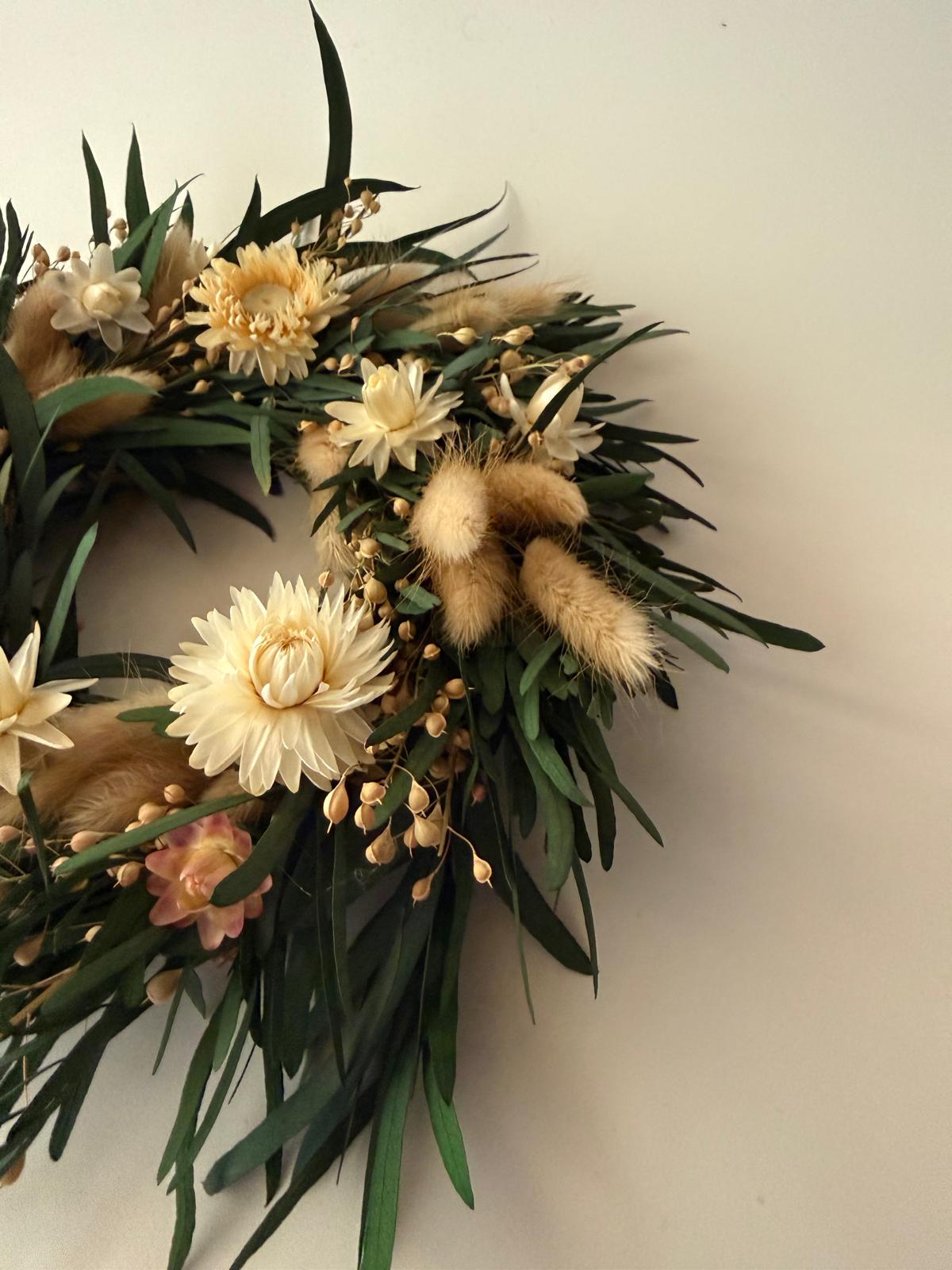 A dried floral wreath featuring cream-colored flowers, green leaves, and fluffy beige grasses, hanging against a plain off-white wall.