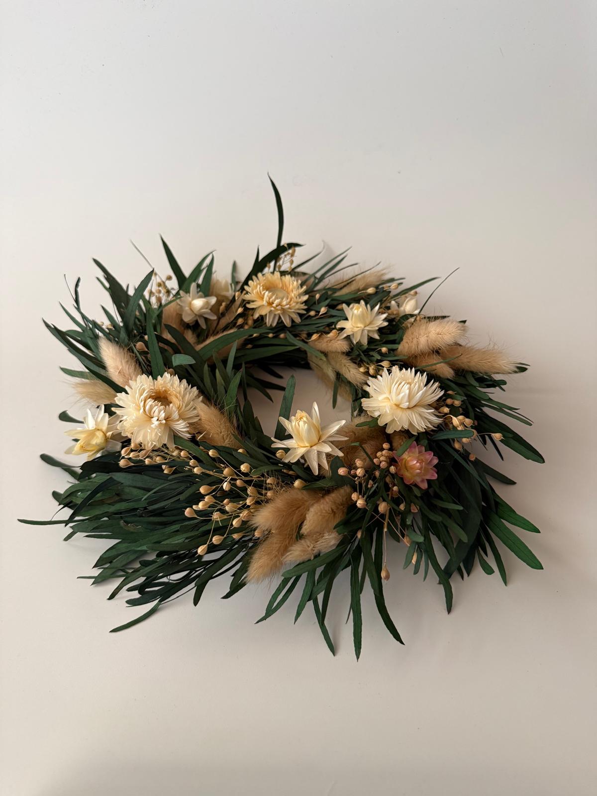 A dried flower wreath featuring cream-colored flowers, green leaves, and fluffy beige pampas grass, displayed against a plain off-white background.