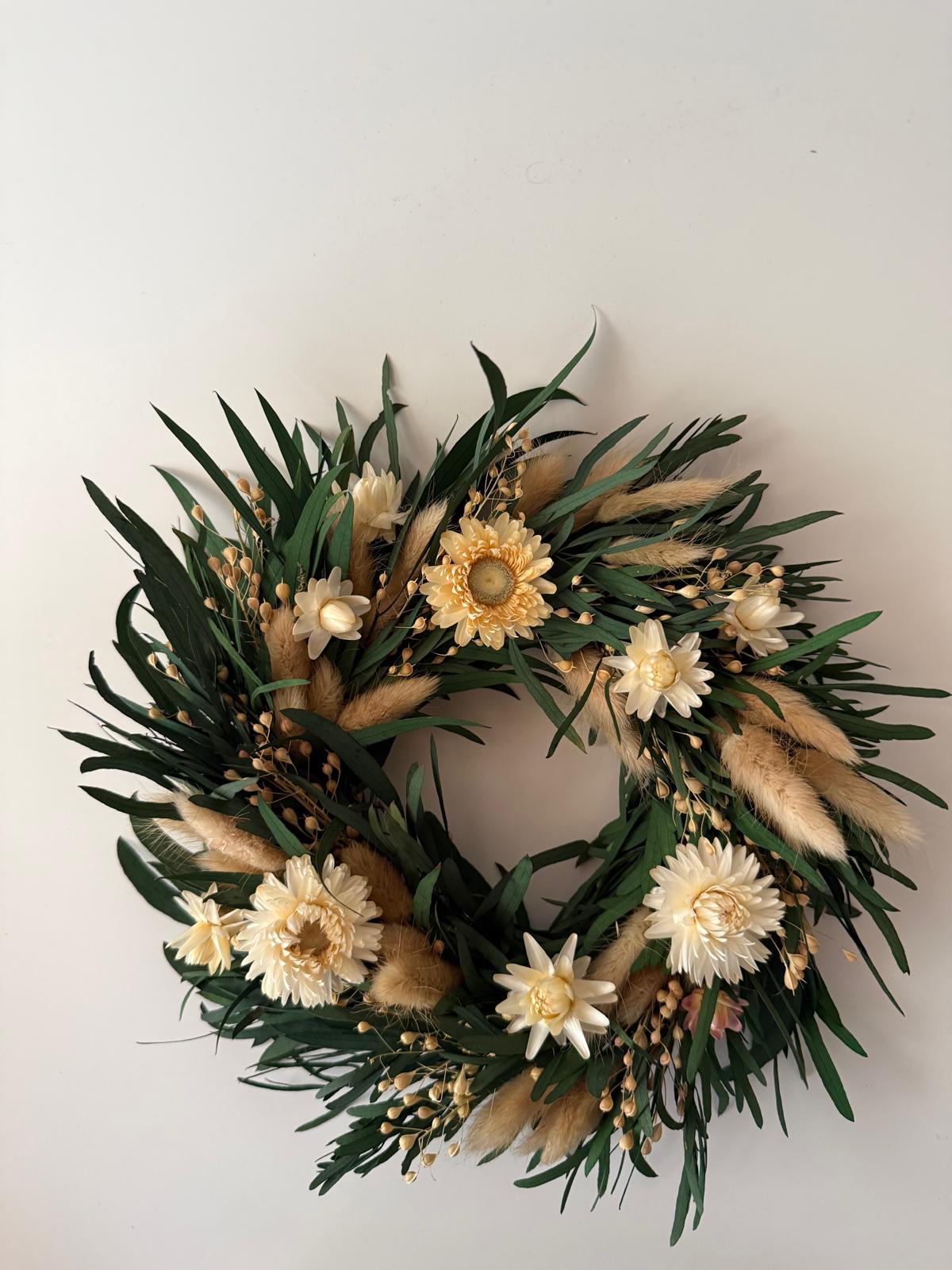 A dried floral wreath featuring green leaves, cream and yellow flowers, and fluffy beige pampas grass, displayed against a plain off-white wall.