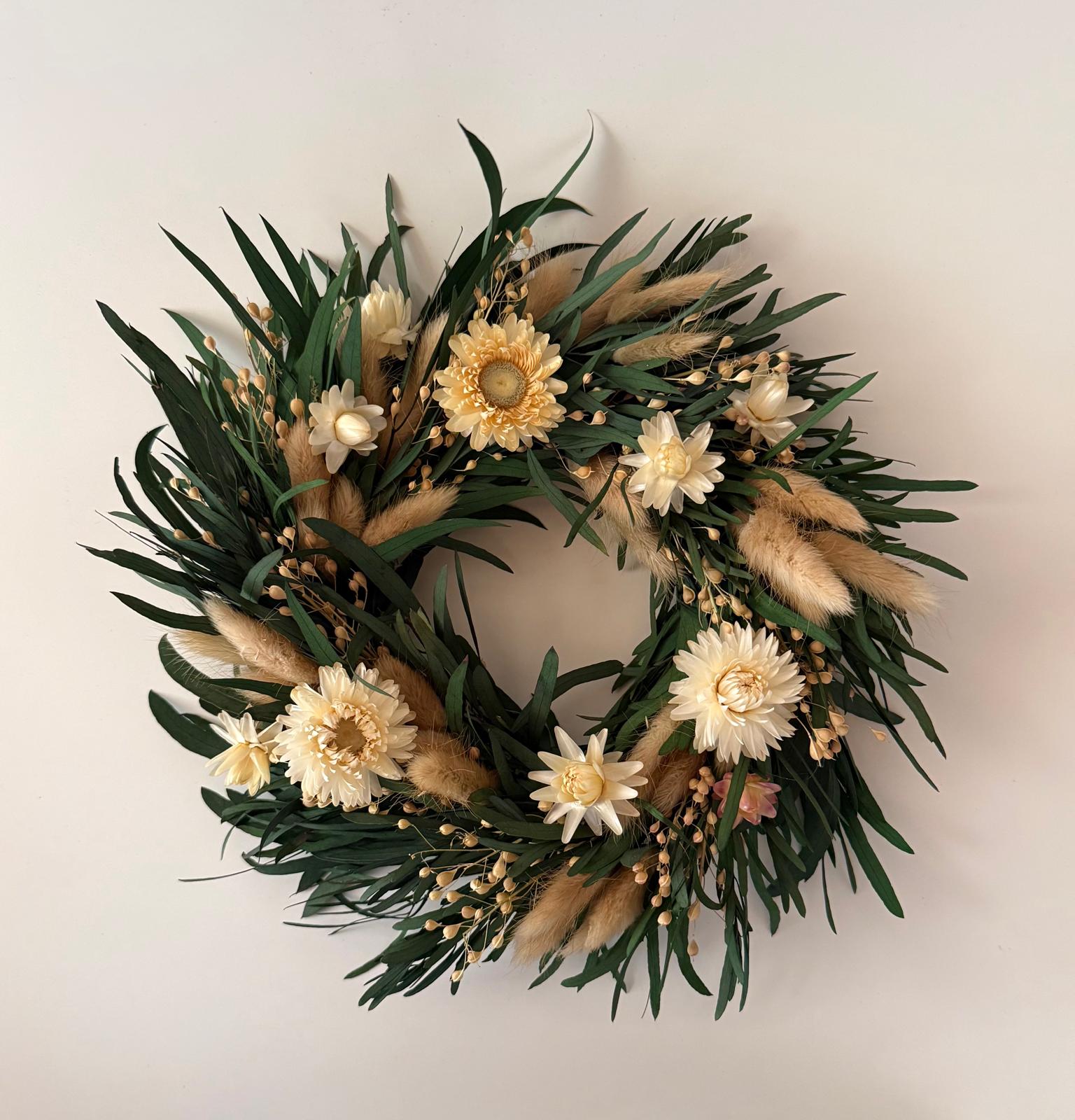 A dried floral wreath featuring green leaves, cream and yellow flowers, and fluffy beige pampas grass, displayed against a plain off-white wall.