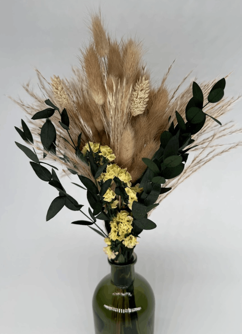 Dried flower arrangement with pampas grass, bunny tails, yellow flowers, and eucalyptus in a green bottle vase on a white background