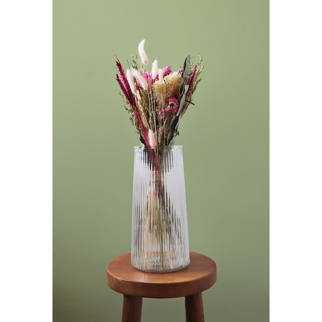Clear glass vase with dried flowers on a wooden stool against a green background