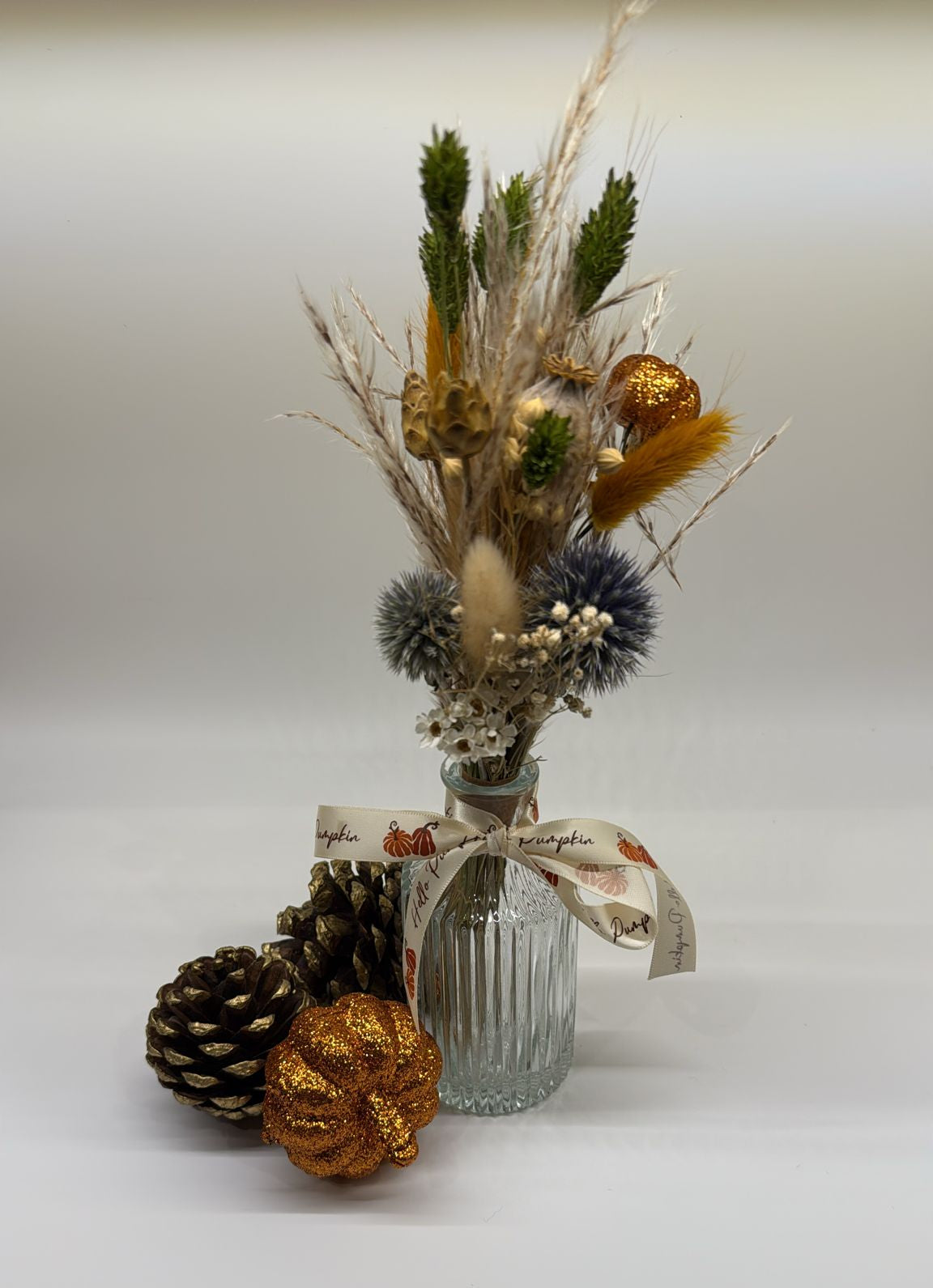 Decorative arrangement with a glass vase, dried flowers, pinecones, and a gold ornament on a white background.