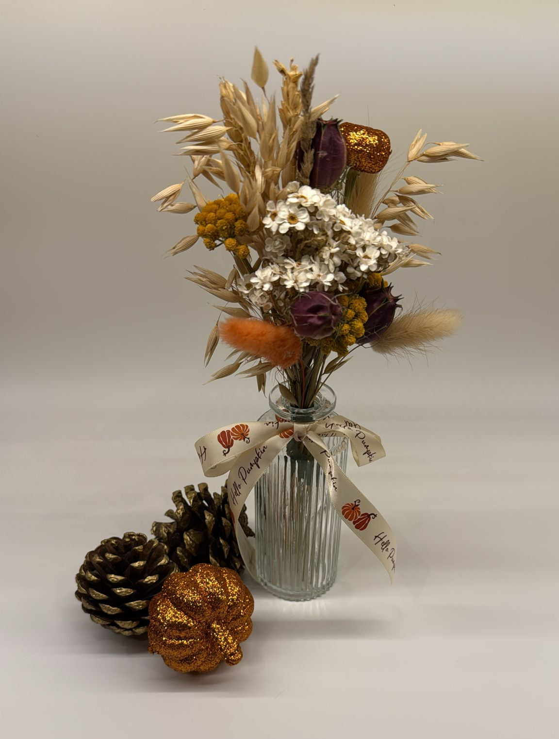 Decorative arrangement with dried flowers, pinecones, and a small pumpkin in a glass vase on a white background.