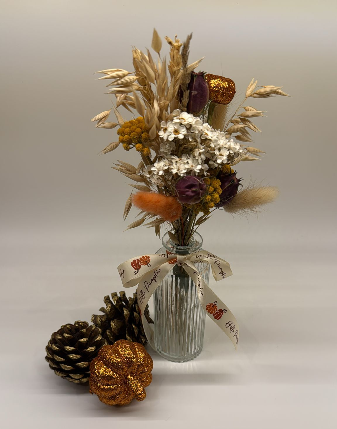 Decorative arrangement of dried flowers in a glass vase with decorative ribbons, accompanied by pinecones and a small pumpkin on a light background.