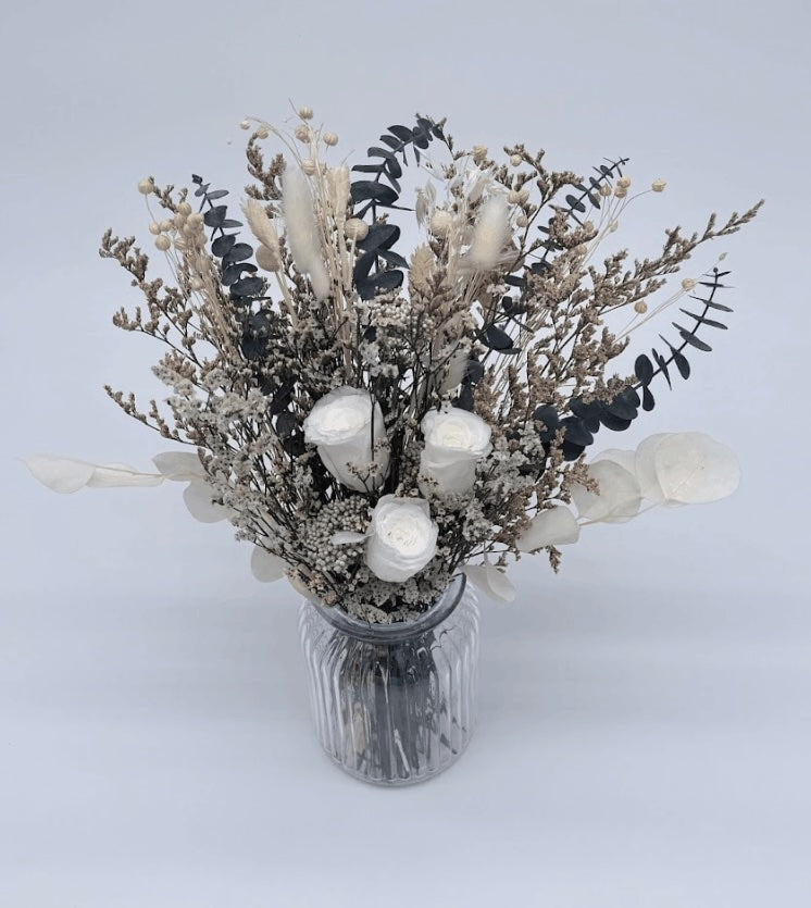 A  bouquet of white roses, fluffy bunny tails, globe amaranths, and icy-textured fillers in soft ivory tones with silvery foliage and deep eucalyptus greens placed into a decorative vase on a wooden surface against a plain background