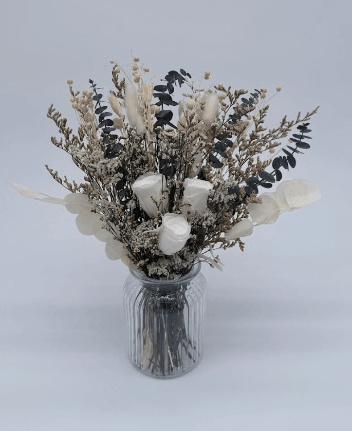 A  bouquet of white roses, fluffy bunny tails, globe amaranths, and icy-textured fillers in soft ivory tones with silvery foliage and deep eucalyptus greens placed into a decorative vase on a wooden surface against a plain background